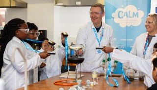 Two adults and multiple schoolchildren use devices to monitor air quality while some sausages are cooked using gas
