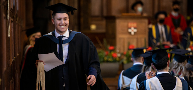 A smiling student walks through the Great Hall in his gap and gown, holding his degree certificate.