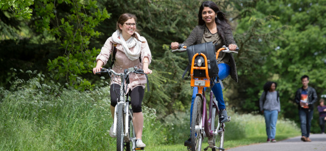 Two students riding bikes against some tall grass.