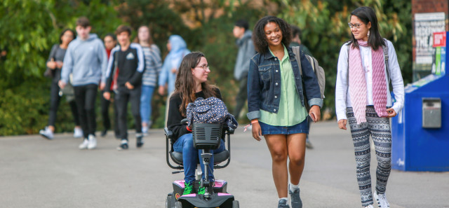 Three female students travel to their lecture. One is in an electric scooter, talking to her two friends as they walk.